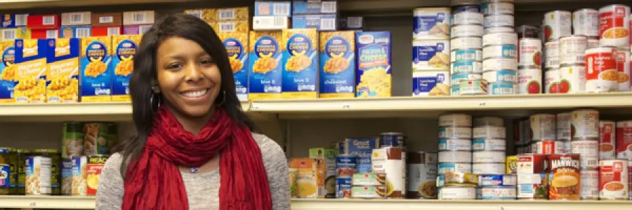 A lady in a grey long sleeve shirt and red scarf holds canned goods while standing in front of shelves of more canned goods.
